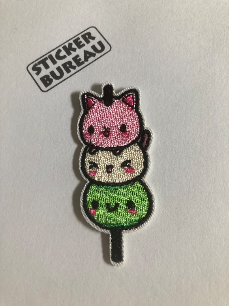 May include: A cute embroidered patch featuring a stack of three green, white, and pink mochi with faces, on a stick.