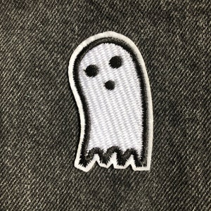 Könnte beinhalten: Ein weißer, geisterförmiger Aufnäher mit schwarzem Umriss und drei schwarzen Punkten als Augen und Mund. Der Aufnäher hat einen gezackten unteren Rand und liegt auf einem dunkelgrauen, strukturierten Hintergrund.