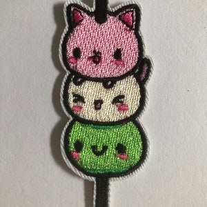 Puede incluir: Un parche bordado con tres caras de gato kawaii apiladas en un palo. El gato de arriba es rosa con ojos negros y una nariz negra. El gato del medio es blanco con ojos negros y una nariz negra. El gato de abajo es verde con ojos negros y una nariz negra.