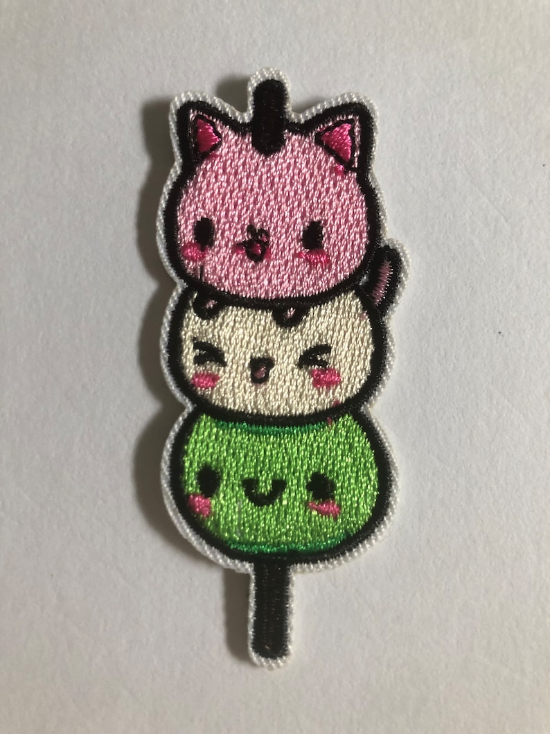 May include: An embroidered patch featuring three stacked kawaii cat faces on a stick. The top cat is pink with black eyes and a black nose. The middle cat is white with black eyes and a black nose. The bottom cat is green with black eyes and a black nose.