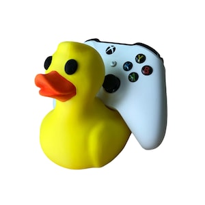 May include: A yellow rubber duck with black eyes and an orange beak is positioned next to a white Xbox controller. The controller has black buttons and colored action buttons.