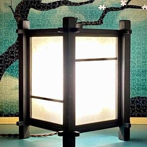 Puede incluir: Una linterna de madera negra con paneles de papel blanco, iluminada desde dentro. La linterna tiene forma cuadrada y está sostenida por cuatro patas. El fondo presenta un diseño de árbol y un rompecabezas.