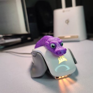 May include: A purple snake-shaped decorative item rests on a white computer mouse. The snake has green eyes and a detailed, textured design. The mouse has a glowing logo and a sleek, modern design, suggesting a gaming accessory.