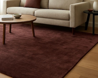 May include: A living room scene featuring a beige sofa with a burgundy throw pillow. A round wooden coffee table sits on a burgundy rug. An abstract art print hangs above the sofa, and a small side table with a lamp is visible.