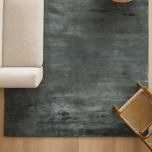 Puede incluir: Vista aérea de un salón con una gran alfombra verde oscuro. A la izquierda, un sofá beige con un cojín gris. En el centro, una mesa de centro redonda de madera con tapa de mármol y un cuenco pequeño. A la derecha, una silla de mimbre.