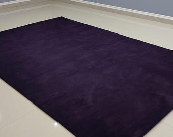 May include: A rectangular, plush, dark purple rug. The rug is the focal point, filling most of the frame. The rug's texture appears soft and inviting. The background features a light-colored floor and a gray wall.