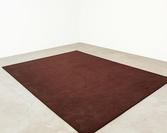 May include: A rectangular, solid brown rug. The rug is placed on a light-colored concrete floor, with white walls in the background. The rug's color is a deep, rich shade of brown, and it appears to be made of a soft material.