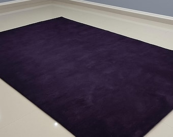 May include: A rectangular, plush, dark purple rug. The rug is the focal point, filling most of the frame. The rug's texture appears soft and inviting. The background features a light-colored floor and a gray wall.