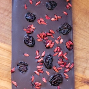 May include: A rectangular dark chocolate bar with dried berries and red fruit pieces scattered across the top. The chocolate is a deep brown colour, and the toppings are visible. The bar is on a wooden surface.