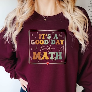May include: Burgundy sweatshirt with the text "It's a good day to do math" in a colorful, handwritten style. The text is surrounded by a white box with math symbols.