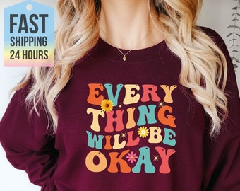 May include: A maroon sweatshirt with the text "EVERYTHING WILL BE OKAY" in colorful retro font with flowers and sparkles.