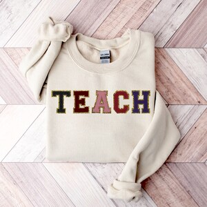May include: A beige sweatshirt with the word "TEACH" in colorful block letters with gold glitter accents.