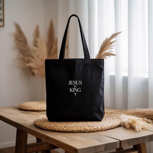 A black tote bag with the text "JESUS KING" printed on it, placed on a woven placemat on a wooden table.