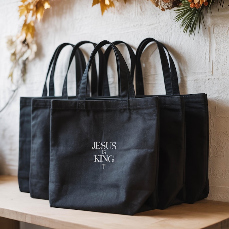 A row of black tote bags with the text "JESUS IS KING" printed on them, placed on a wooden surface against a white wall with some decorative elements.