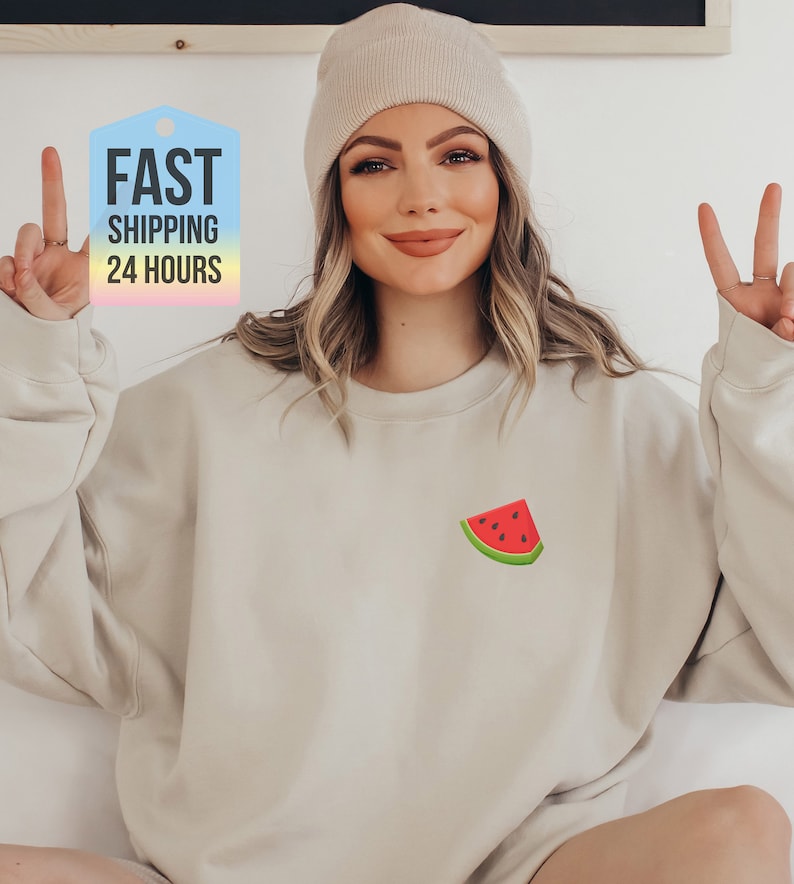 May include: A light beige sweatshirt with a small watermelon graphic on the front. The sweatshirt has a crew neck and long sleeves.