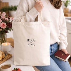 A woman wearing a white sweater and jeans is sitting at a kitchen counter, holding a white tote bag with the text "JESUS IS KING" printed on it. The counter also has a bowl of strawberries, a candle, and a book.