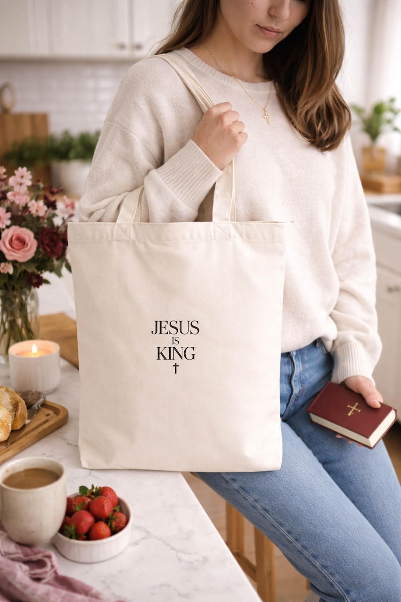A woman wearing a white sweater and jeans is sitting at a kitchen counter, holding a white tote bag with the text "JESUS IS KING" printed on it. The counter also has a bowl of strawberries, a candle, and a book.