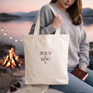 A woman sits by a campfire, holding a white tote bag with the text "JESUS IS KING" printed on it.