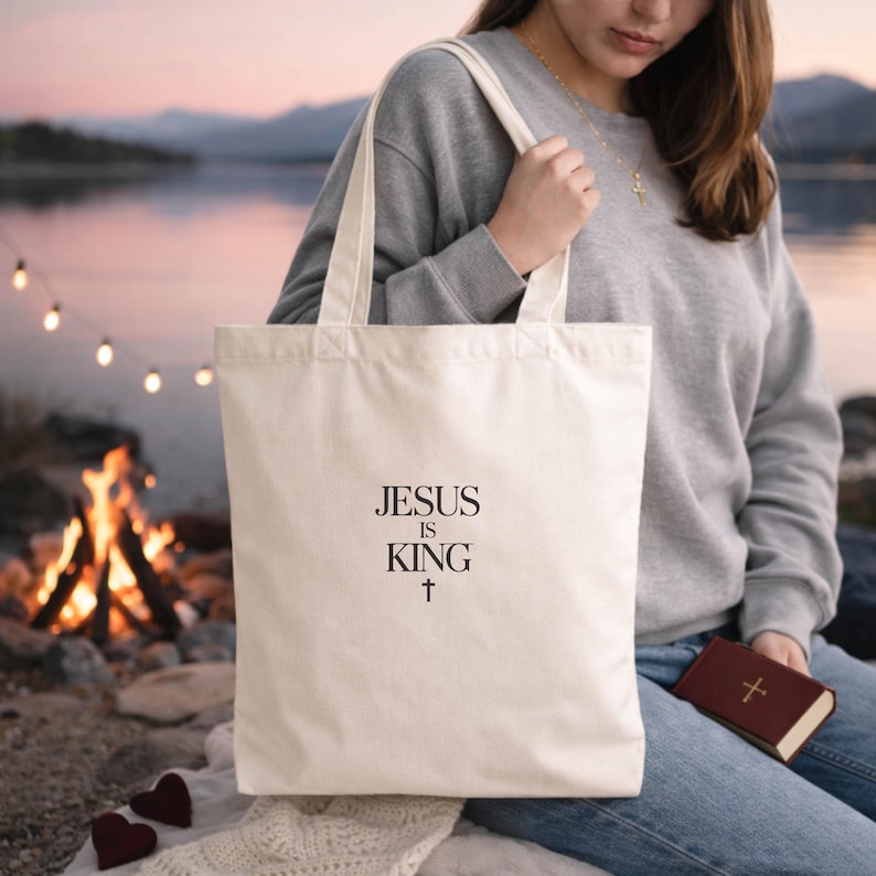 A woman sits by a campfire, holding a white tote bag with the text "JESUS IS KING" printed on it.