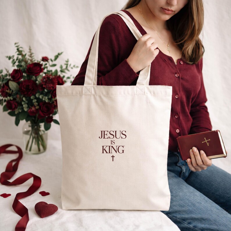 A woman in a burgundy sweater sits on a white surface, holding a white tote bag with the text "JESUS IS KING" printed on it. Next to her is a red book and a vase filled with red roses.