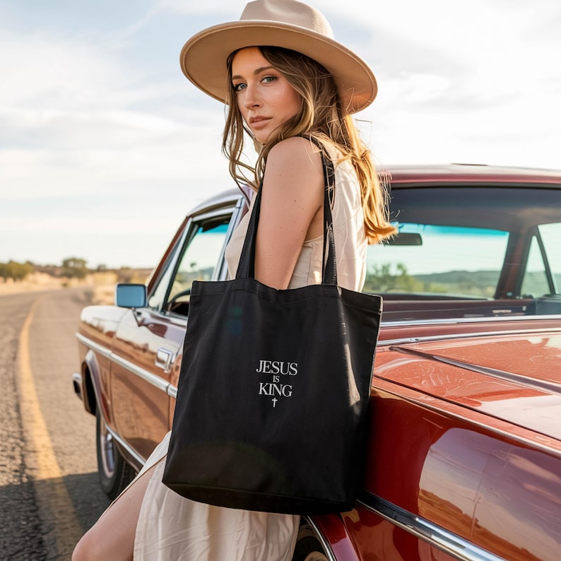 A woman in a hat stands next to a red car, holding a black tote bag with the text "JESUS IS KING" printed on it.