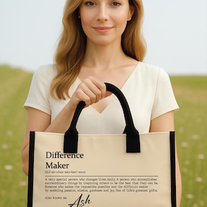 May include: A beige tote bag with black accents and handles. The bag features the text "Difference Maker" and a definition, with the name "Ash" below. The woman in the image is holding the bag.