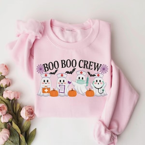 May include: A light pink crewneck sweatshirt featuring a graphic of four ghosts dressed as medical professionals with the text "BOO BOO CREW" above them. The ghosts are accompanied by orange pumpkins, purple spiderwebs, and black bats.