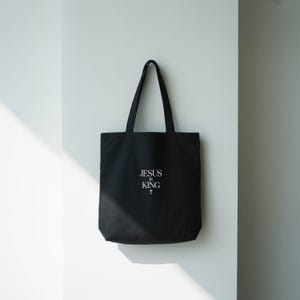 A black tote bag with the text "JESUS IS KING" printed on it, hanging on a white wall.
