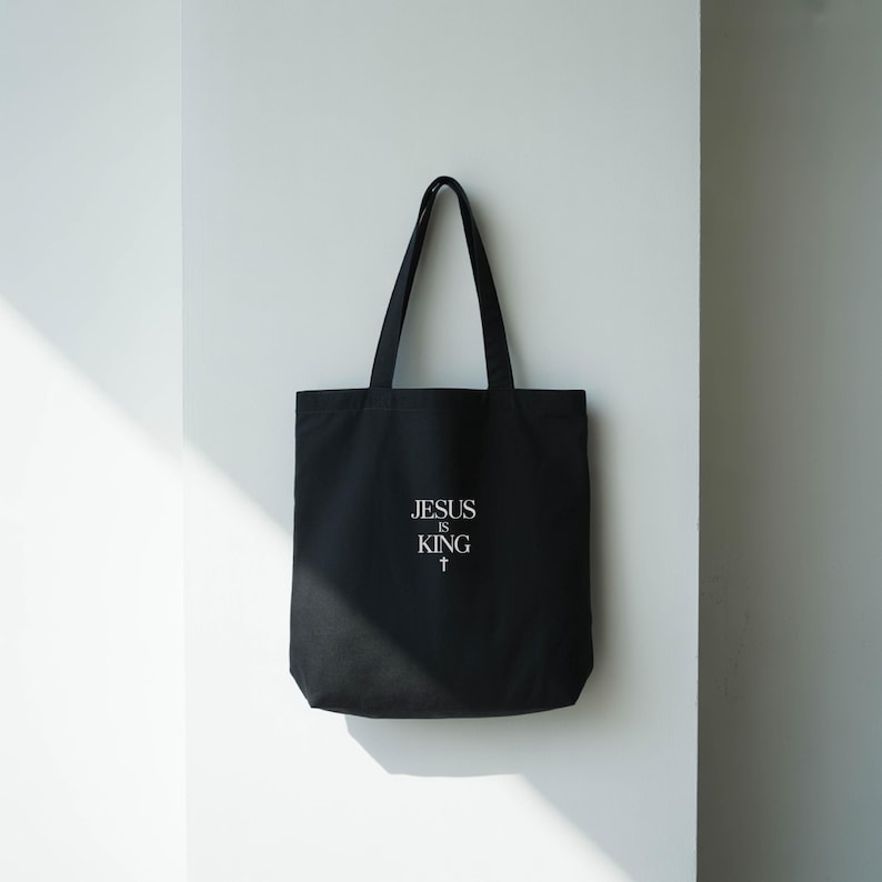 A black tote bag with the text "JESUS IS KING" printed on it, hanging on a white wall.