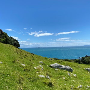 Puede incluir: Una ladera cubierta de hierba con vistas a un océano azul con algunas pequeñas islas en la distancia. El cielo es de un azul brillante con nubes blancas.