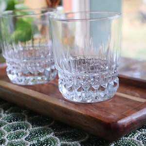 May include: Two clear glass tumblers with a diamond-cut pattern sit on a wooden serving tray. The tray has a light wood inlay and is made of dark wood.