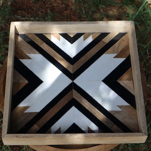 Peut inclure: Un plateau carré en bois avec un motif géométrique. Le motif est composé de pièces de bois noir, blanc et brun disposées en forme de flèches. Le plateau est parfait pour servir des collations ou des boissons.