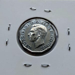 May include: A silver-colored coin featuring a profile portrait of King George VI facing left. The inscription GEORGIVS VI D:G:REX ET IND:IMP: is embossed around the edge. The coin is secured within a square white cardboard holder with staples.