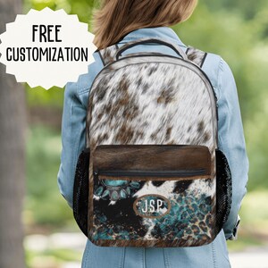 May include: A backpack featuring a white and brown cowhide print on the top section and a dark brown cowhide print on the bottom pocket. The front pocket displays a turquoise gemstone graphic, leopard print accents, and an oval patch with the text JSP.