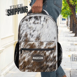 May include: A brown and white cowhide print backpack with a black zipper and a black mesh pocket on each side. The backpack has a black patch with the word "MADISON" in white lettering.