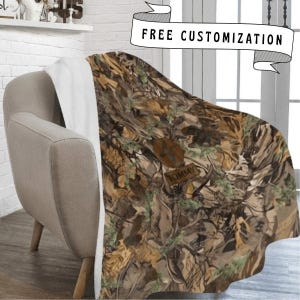 May include: A plush throw blanket featuring a brown, tan, and green camouflage pattern with a white underside. The blanket displays a circular emblem with a duck silhouette and a rectangular patch with the name TOMMY printed in black text.