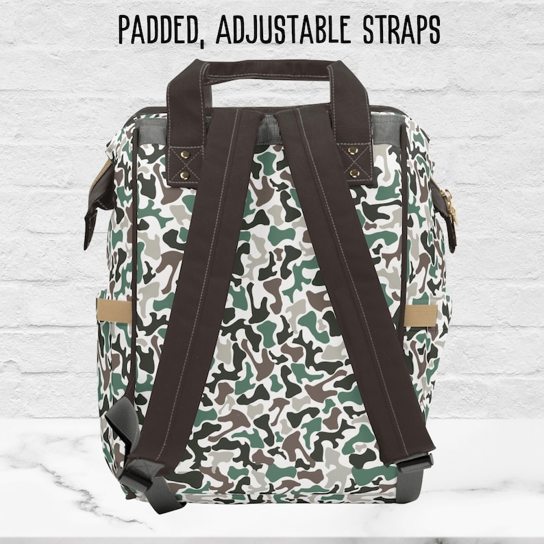 The back side of our camouflage-patterned backpack with padded, adjustable straps. The backpack is primarily green, white, and brown, with a dark brown canvas strap. The text 'PADDED, ADJUSTABLE STRAPS' is displayed at the top.