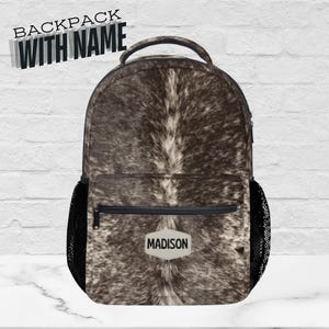 May include: A backpack with a brown and white cowhide pattern. The backpack has a top handle, a front pocket, and two side mesh pockets. The name "MADISON" is displayed on a white label. The text "BACKPACK WITH NAME" is at the top.