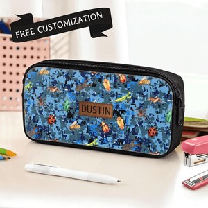 May include: A rectangular black pencil case featuring a blue digital camouflage pattern with various insect illustrations and a brown rectangular patch in the centre with the name DUSTIN printed on it. A black banner above reads FREE CUSTOMIZATION.