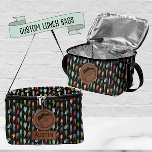 May include: Two black insulated lunch bags with a fishing lure pattern. One bag is open, revealing a silver insulated interior. Both bags have a brown leather patch with a fish logo. One bag has the name "Austin" on it. The text "CUSTOM LUNCH BAGS" is above the bags.