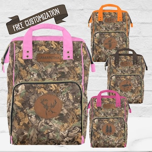 May include: Four camouflage backpacks with pink, orange, brown, and pink trim. Each backpack has a personalized name tag and a different design on the front. The designs include a deer, a duck, a fish, and a pine tree.