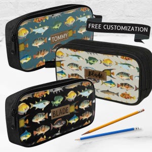 Peut inclure: Trois trousses rectangulaires avec fermetures éclair et bordures noires, présentant un motif de poissons répété sur des fonds bleu, beige et noir. Chaque trousse possède un écusson rectangulaire central avec les noms TOMMY ou BLADE.