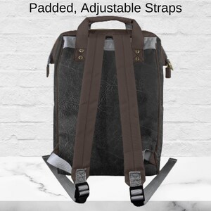 May include: A black and brown backpack with padded, adjustable straps. The backpack has a top handle and a zipper closure. The straps are brown, and the top of the bag has a gray accent. The text "Padded, Adjustable Straps" is at the top.