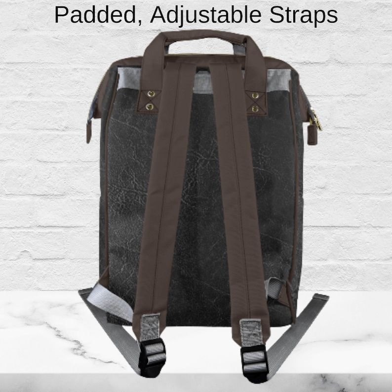 May include: A black and brown backpack with padded, adjustable straps. The backpack has a top handle and a zipper closure. The straps are brown, and the top of the bag has a gray accent. The text "Padded, Adjustable Straps" is at the top.