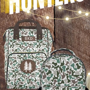 May include: Three camouflage-patterned bags with brown leather accents. The bags feature different embroidered designs, including a pair of trees, a duck, and a deer. The bags are labeled with the initials "BKD".  Valentine's Day gifts for hunters.
