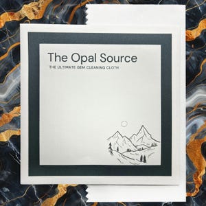 以下が含まれることがあります： 濃い緑色の縁取りのある白いカード。カードの上部に黒で「The Opal Source」というテキストが印刷されています。テキストの下には「The Ultimate Gem Cleaning Cloth」というフレーズがあります。カードの下部には、太陽が空にある山脈の白黒のイラストが描かれています。