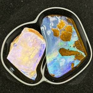 May include: Two pieces of opal, one with a blue and green iridescent sheen and the other with a white and purple iridescent sheen, are set in a black resin background.