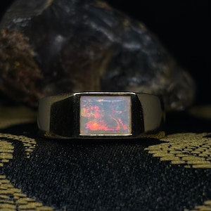 Può includere: Un anello con fascia in oro con una pietra preziosa di opale di forma quadrata. L'opale è di colore bianco latte con bagliori di rosso, arancione e giallo.