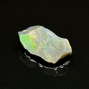 May include: A rough, white opal gemstone with iridescent green and blue flashes. The opal is irregularly shaped and has a natural, unpolished finish.