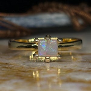 Puede incluir: Un anillo de oro con una piedra de ópalo de forma cuadrada. El ópalo es iridiscente con tonos de azul, verde y blanco.